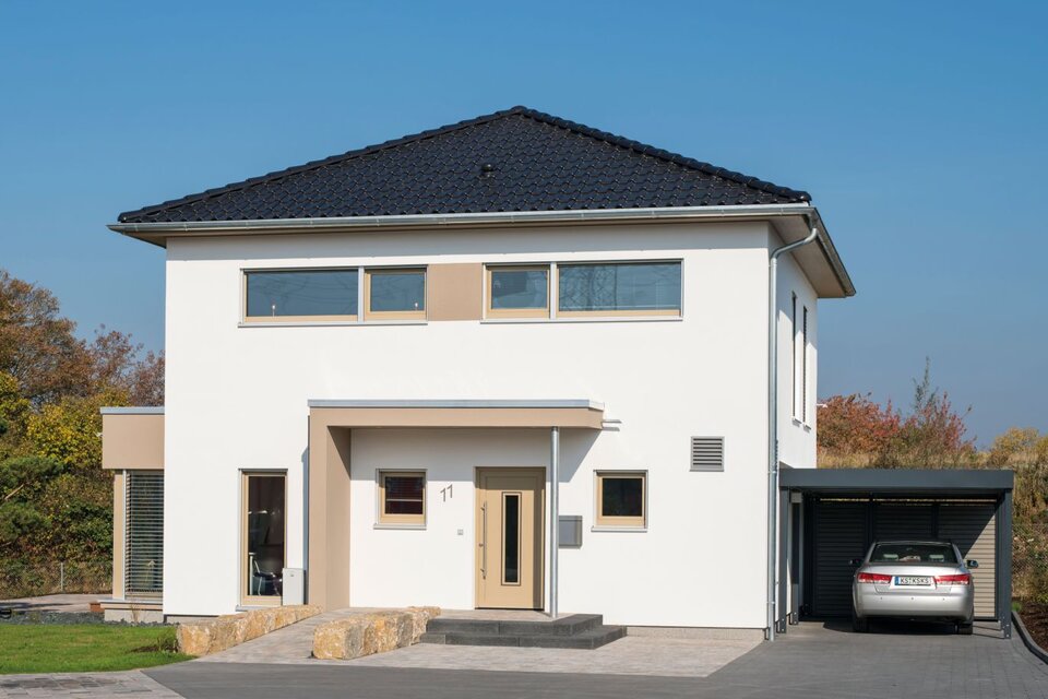 Frontansicht des Musterhauses MEDLEY 300 B in Kassel mit Walmdach, heller Putzfassade und Carport mit geparktem Auto.