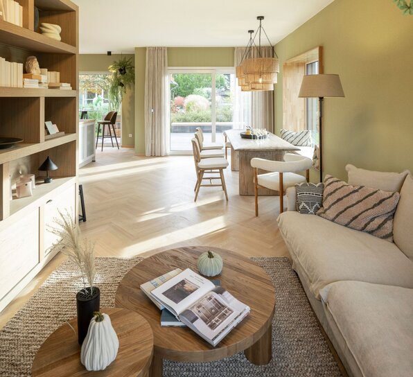 Wohn- und Essbereich im Musterhaus JUNO 300 in Mülheim-Kärlich mit hellen Holzböden, gemütlichem Sofa, rundem Couchtisch und großem Esstisch am Fenster.