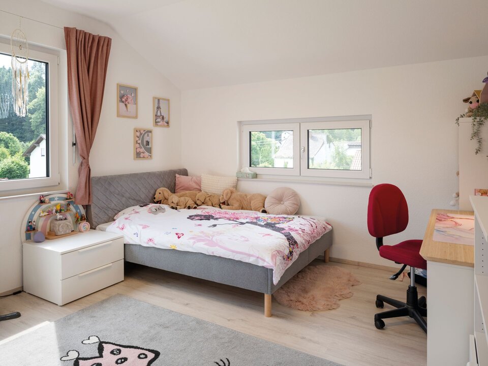 Kinderzimmer mit Bett, Schreibtisch und Fenster. Warme Farben, Teppich und rosa Vorhänge sorgen für eine gemütliche Atmosphäre.