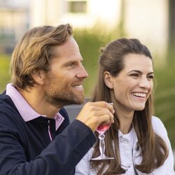 Eine Gruppe von Freunden sitzt im Garten an einem gedeckten Tisch und stößt mit Gläsern an. Auf dem Tisch stehen Salate, Getränke und Schalen mit Speisen, im Hintergrund sind Pflanzen und Grün zu sehen.