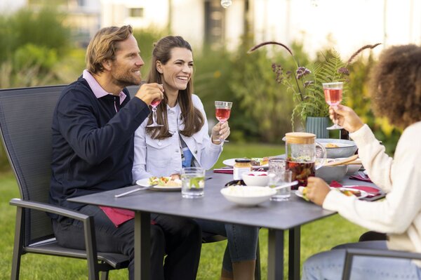 Eine Gruppe von Freunden sitzt im Garten an einem gedeckten Tisch und stößt mit Gläsern an. Auf dem Tisch stehen Salate, Getränke und Schalen mit Speisen, im Hintergrund sind Pflanzen und Grün zu sehen.