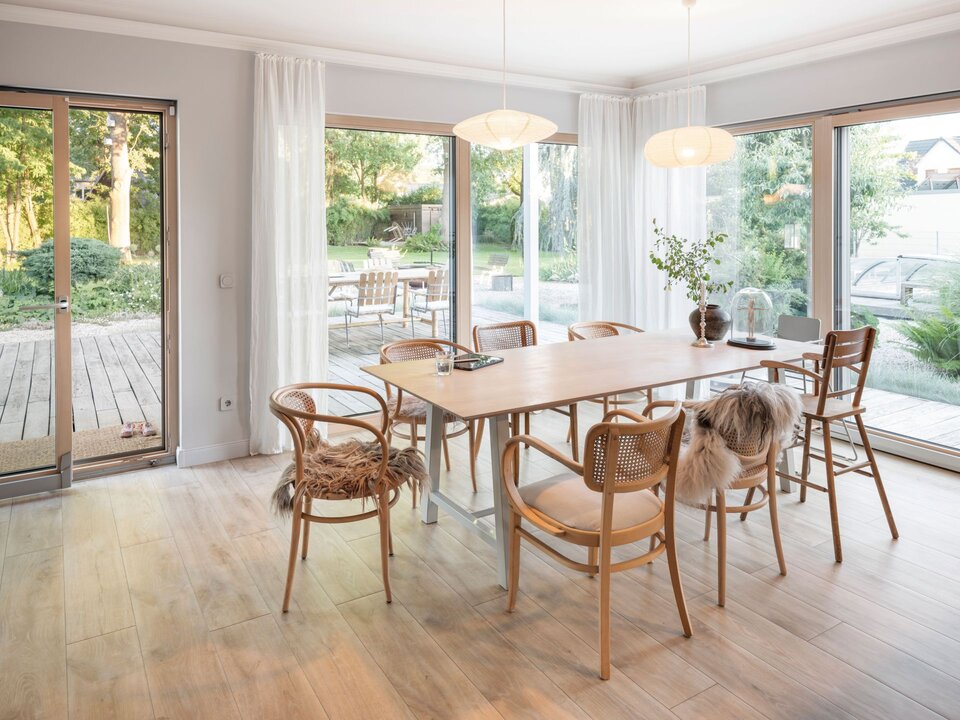 Essbereich mit großem Holztisch, Stühlen aus Naturmaterial und breiter Fensterfront. Helle Vorhänge, Holzfußboden und Blick auf Terrasse und Garten sorgen für eine freundliche Raumatmosphäre.
