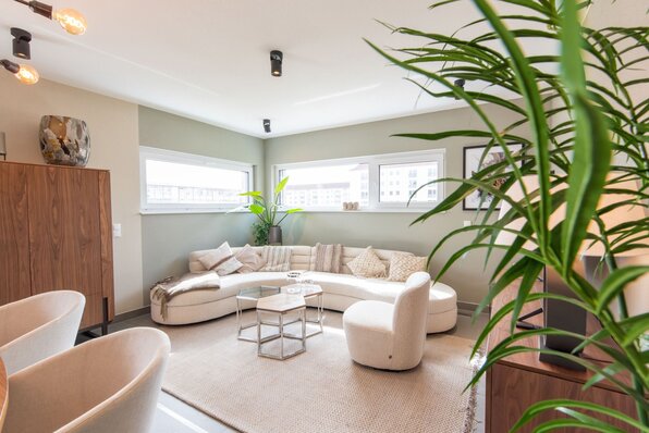 Helles Wohnzimmer im FingerHaus Musterhaus TALO in Babenhausen mit gemütlichem weißen Rundsofa, modernen Beistelltischen und großen Fenstern, die viel Tageslicht hereinlassen. Stilvolle Dekoration mit Pflanzen und Holzmöbeln.