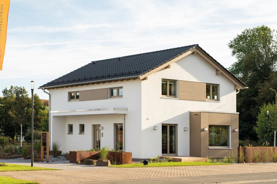 Außenansicht des Finger-Haus Musterhauses SENTO 500 in Frankenberg mit weißer Putzfassade, dunkel gedecktem Satteldach und Holzfassaden-Elementen; davor gepflasterter Weg, bepflanzte Hochbeete, Rasenfläche und Fahnenmasten mit FingerHaus-Flaggen.