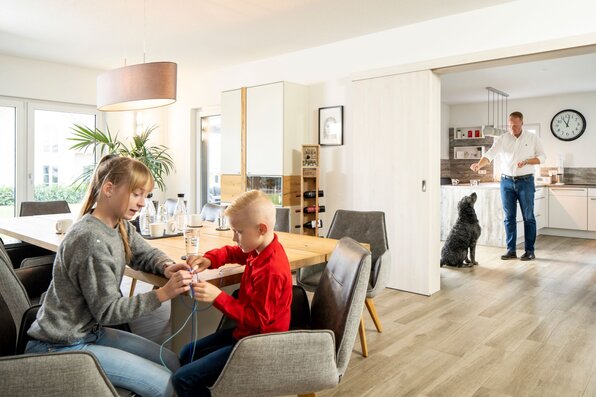 Zwei Kinder basteln am Esstisch, während im Hintergrund ein Mann in der Küche einem Hund ein Leckerli gibt.