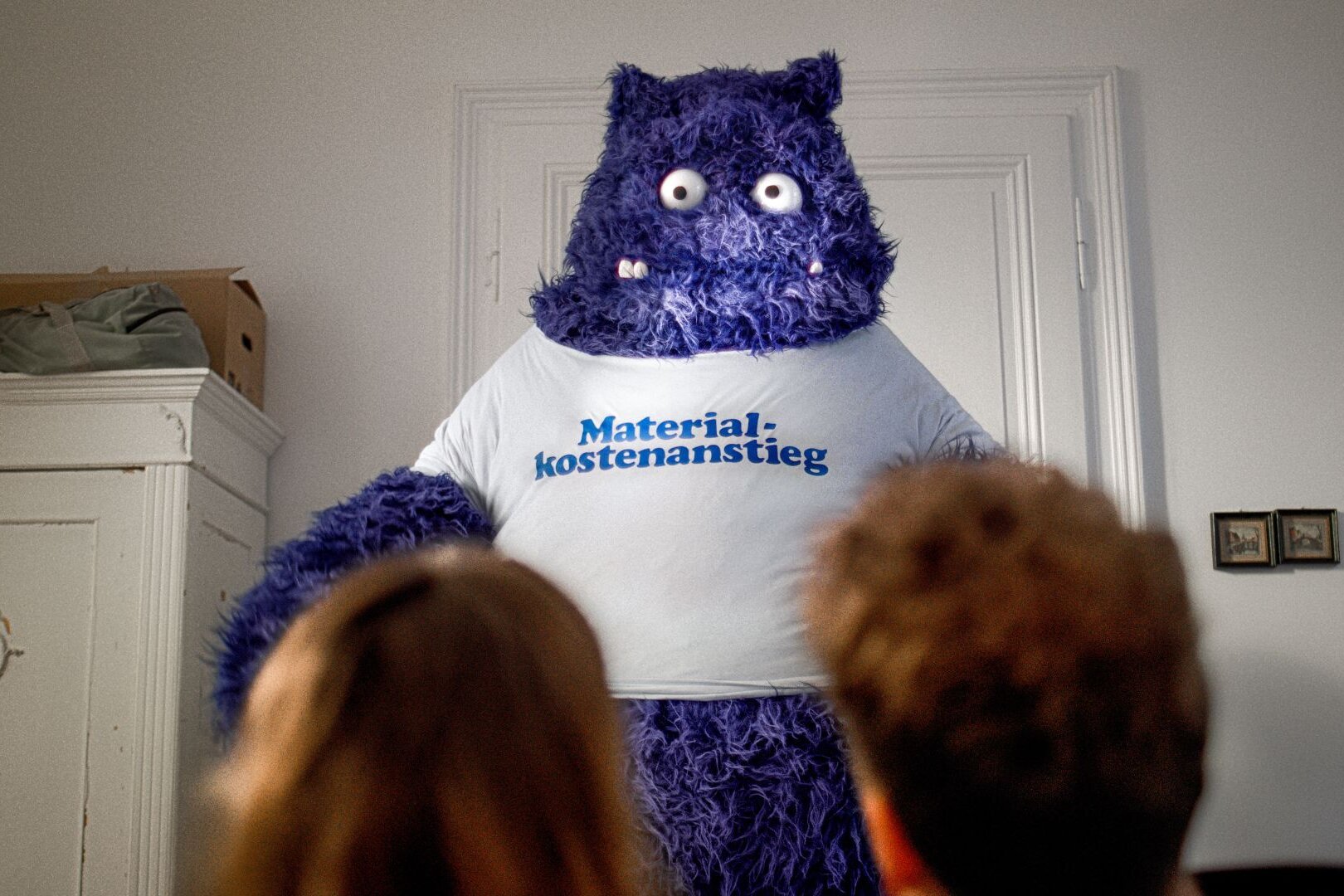 Ein großes blaues Monster mit der Aufschrift „Materialkostenanstieg“ steht in einem Raum und blickt auf zwei Personen, die ihm gegenüber sitzen – Teil der FingerHaus-Kampagne „Das Unerwartete“.