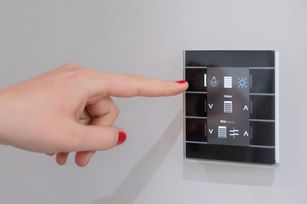 Hand mit rot lackierten Fingernägeln bedient ein schwarzes Touch-Bedienfeld an einer hellgrauen Wand zur Steuerung von Licht und Jalousien.