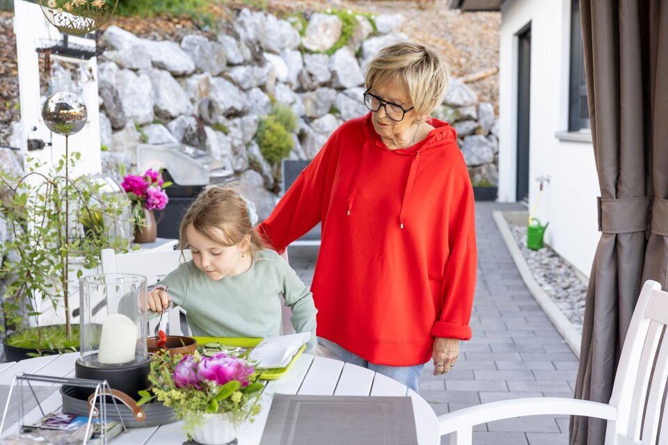 Seniorin begleitet ein Kind am Gartentisch des Kundenhauses NIVO 80 beim gemeinsamen Basteln oder Malen im Außenbereich.