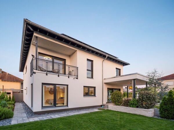 Rückansicht der frei geplanten Stadtvilla mit Balkon, überdachter Terrasse und gepflegtem Garten in der Abenddämmerung.