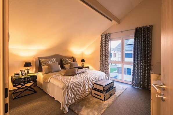 Modernes Schlafzimmer im FingerHaus NEO Musterhaus Günzburg mit großen Fenstern, gemütlicher Beleuchtung und stilvollem Doppelbett.