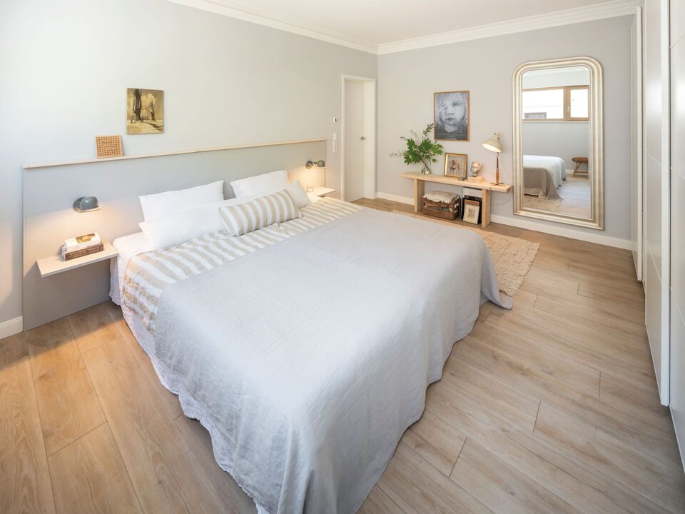 Helles Schlafzimmer mit großem Bett, Holzboden, Wandspiegel und minimalistischer Dekoration in natürlichen Farben.