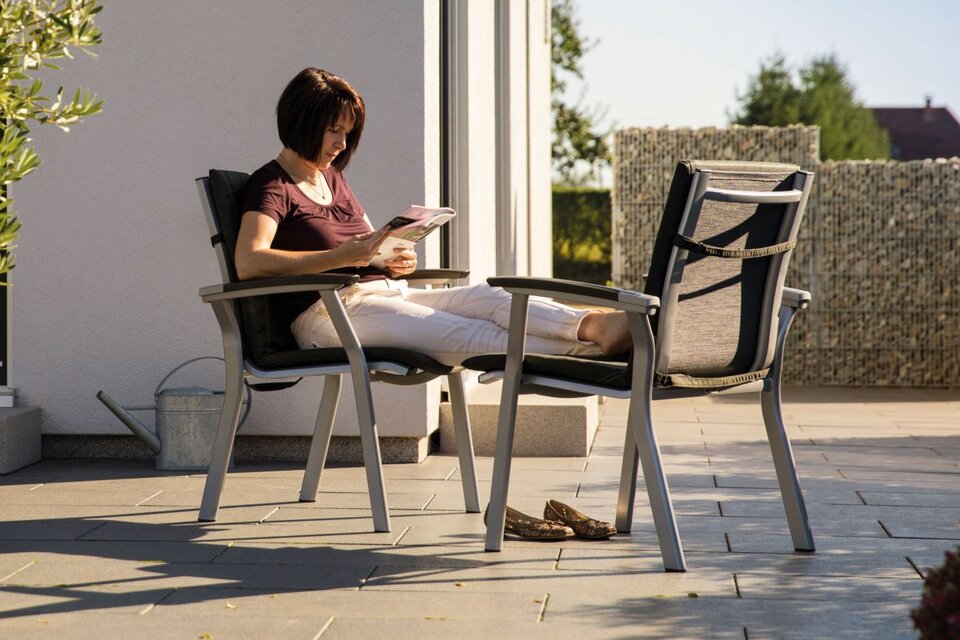Eine Frau sitzt entspannt auf der Terrasse, liest ein Magazin und genießt die Sonne vor ihrem modernen Haus.