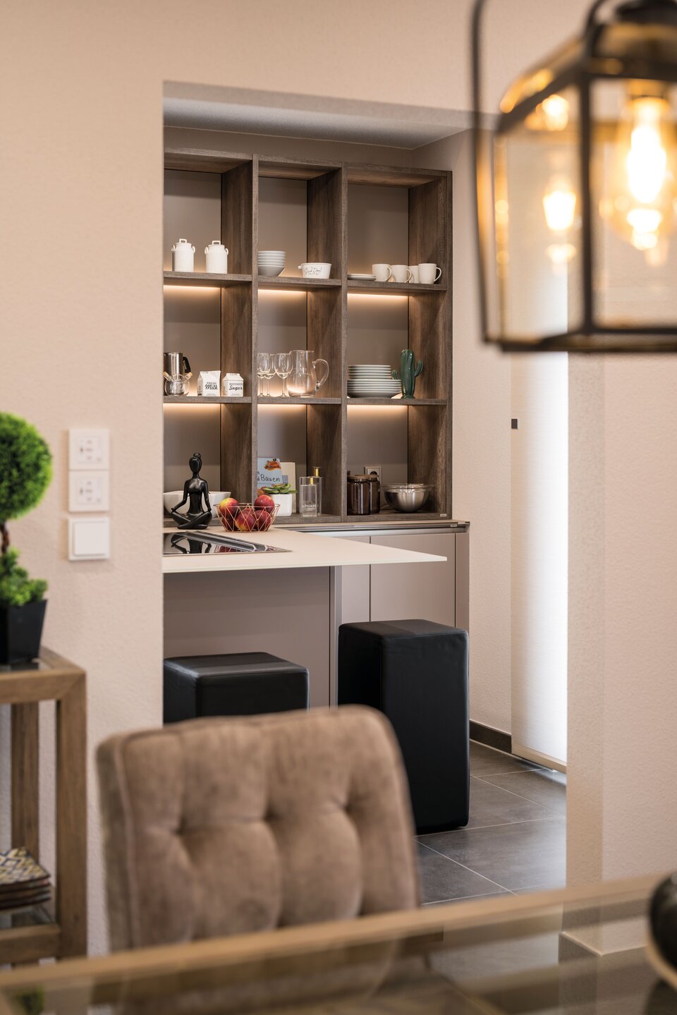 Gemütlicher Küchenbereich mit offenem Holzregal, beleuchteten Fächern und kleiner Arbeitsfläche. Zwei schwarze Hocker stehen darunter, im Vordergrund ein Essbereich mit Glaslampe und gepolstertem Stuhl im Musterhaus UNO Leipzig.
