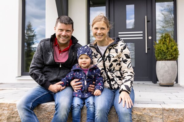Eine Familie sitzt lächelnd vor dem Hauseingang, Eltern mit ihrem kleinen Kind in wetterfester Kleidung.