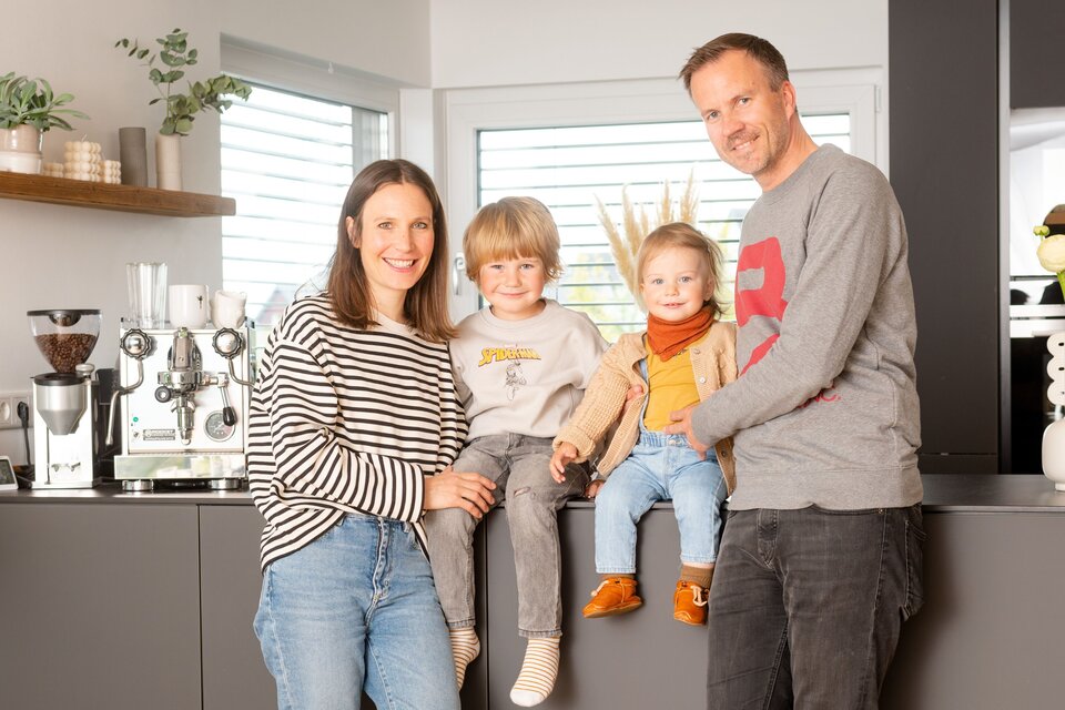 Eine vierköpfige Familie steht und sitzt in der modernen Küche ihres FingerHaus-Hauses. Die Eltern lächeln, während die beiden Kinder auf der Arbeitsplatte sitzen.