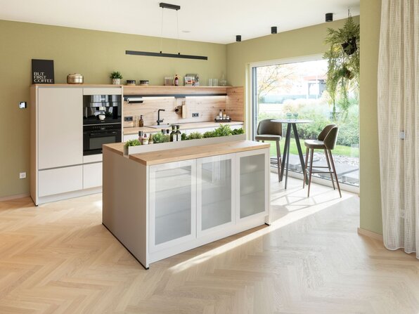 Moderne Küche mit zentraler Kochinsel, weißen Fronten und Holzarbeitsplatte im Musterhaus JUNO Mülheim-Kärlich. Große Fensterfront mit Blick in den Garten, grüne Wandfarbe und integrierte Sitzgelegenheit mit zwei Barhockern.
