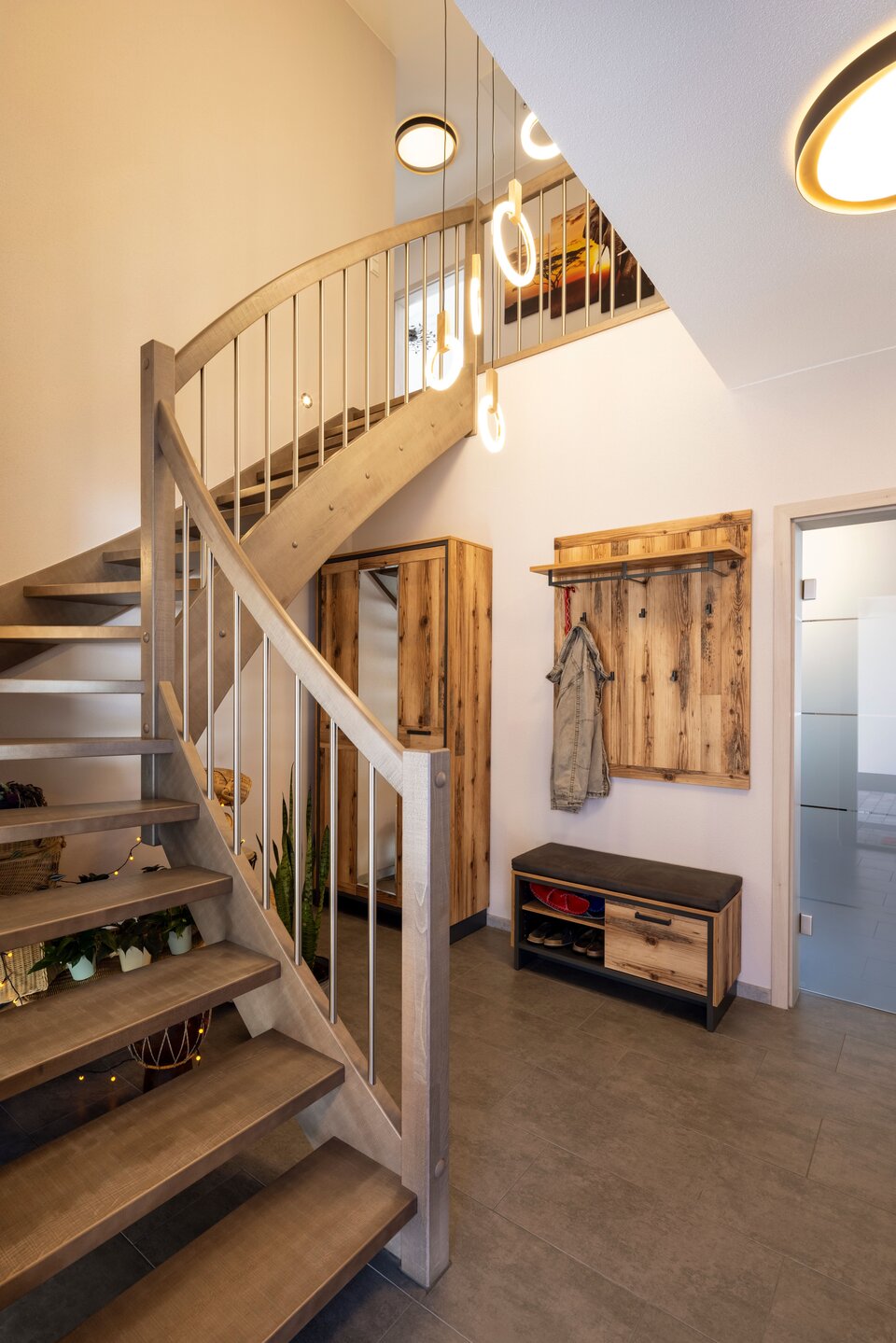 Offene Holztreppe mit geschwungenem Geländer in einem modernen Eingangsbereich. Garderobe und Sitzbank aus Holz, dunkler Boden und Pendelleuchten schaffen ein warmes, einladendes Ambiente.