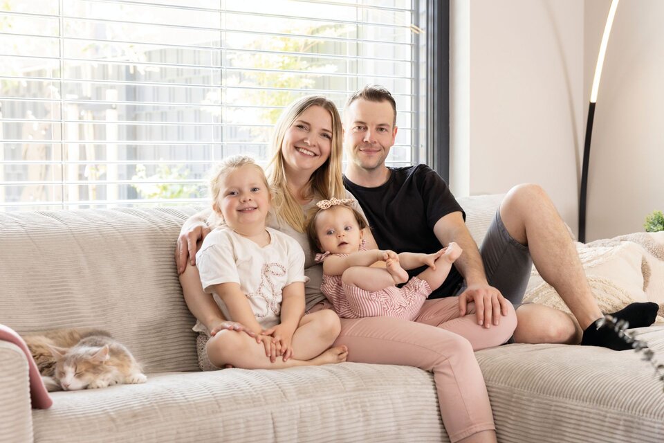 Familie sitzt gemeinsam auf einem Sofa im hellen Wohnzimmer; große Fensterfront, gemütliche Atmosphäre.
