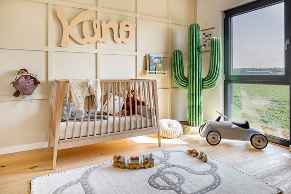 Helles Babyzimmer in einem FingerHaus-Haus mit Holz-Gitterbett, großem Fenster, verspielter Wandgestaltung, Kuscheltieren und Dekoration in warmen Naturtönen.