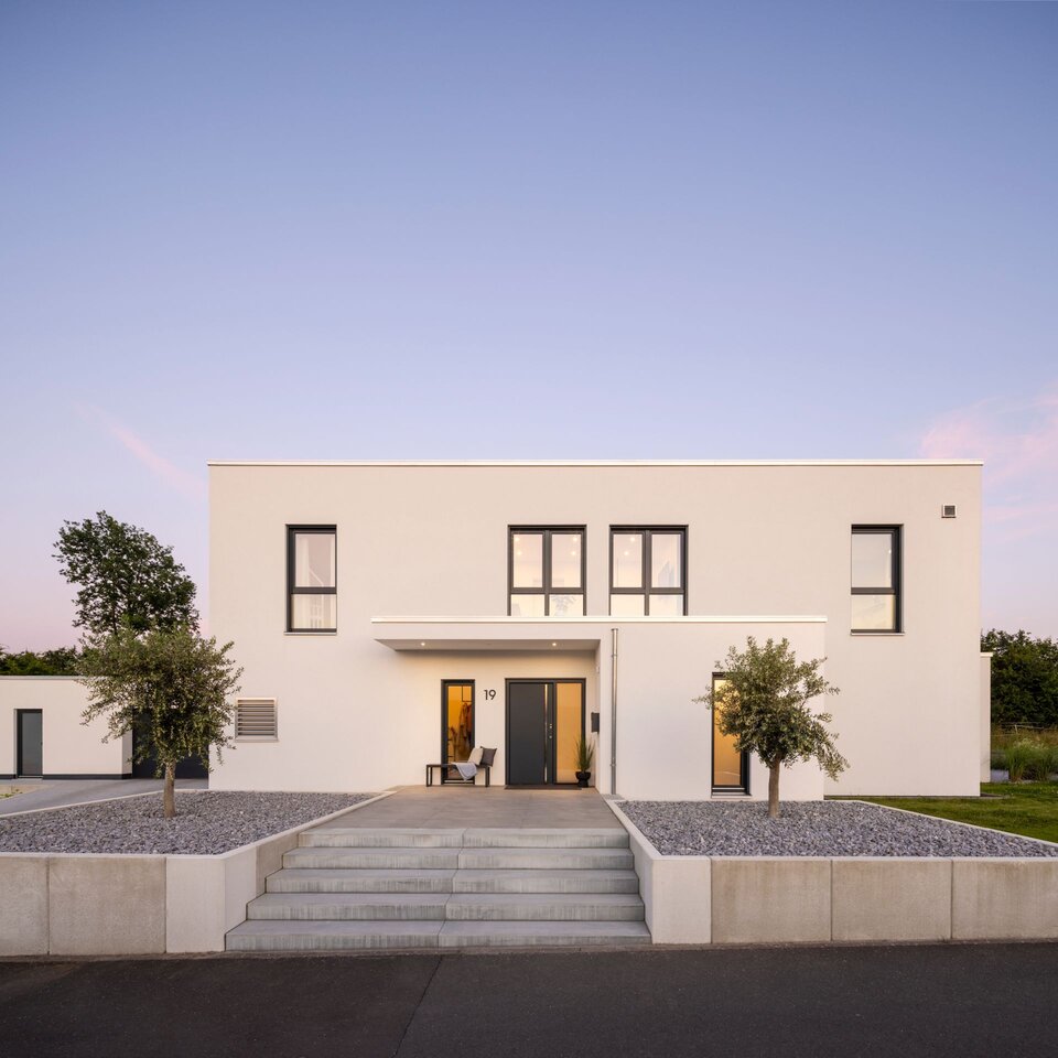 Frontansicht eines modernen FingerHaus im Bauhaus-Stil während der Abenddämmerung. Klare Linien, weiße Fassade und ein breiter Eingangsbereich mit Treppe.