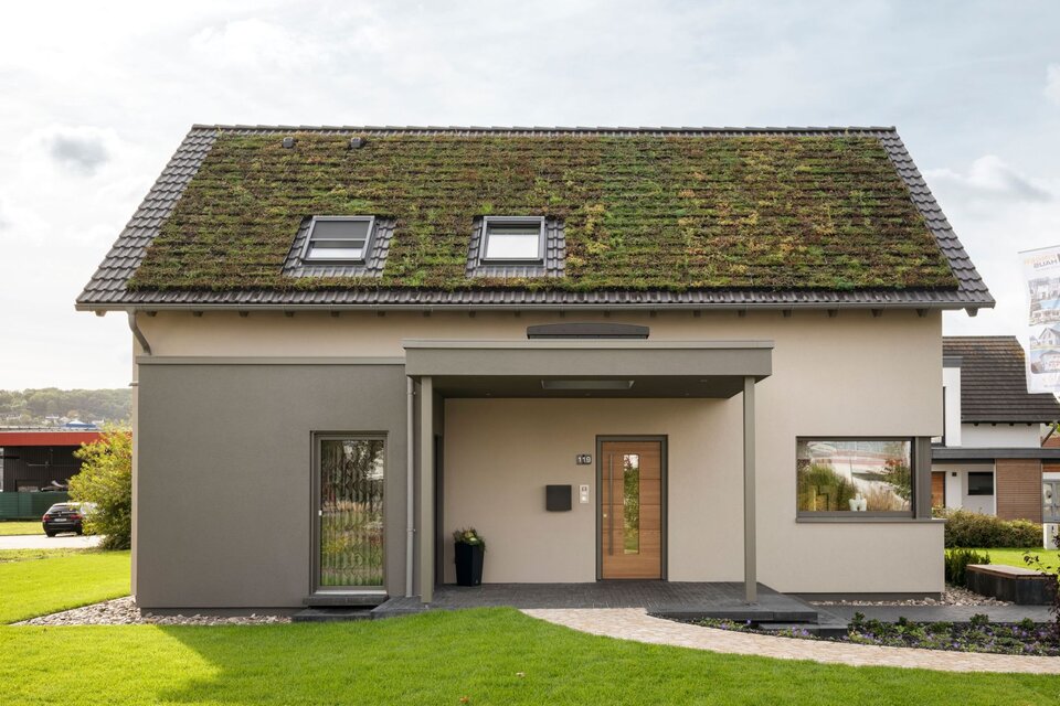 Frontansicht des Musterhauses JUNO 300 in Mülheim-Kärlich mit begrüntem Dach, überdachtem Eingangsbereich und moderner Fassadengestaltung.