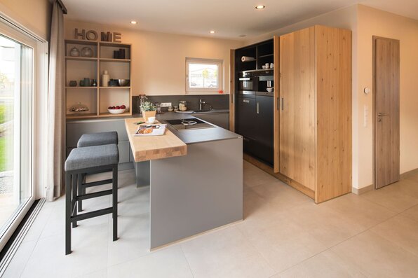 Moderne Küche im FingerHaus NEO Musterhaus Günzburg mit Kochinsel, Holz- und Grauelementen sowie gemütlicher Theke mit Barhockern.