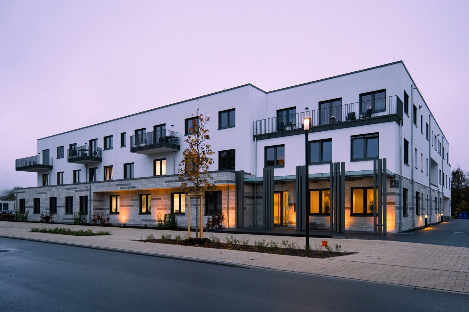 Moderne mehrgeschossige Wohnanlage bei Abendlicht; große Fenster, klare Linien, gehwegnahe Bepflanzung.