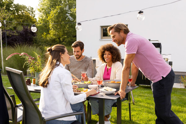 Vier Personen sitzen und stehen an einem Esstisch im Garten neben einem modernen Haus, essen gemeinsam und unterhalten sich, Lichterkette über dem Tisch
