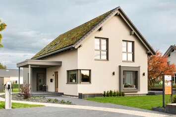 Straßenansicht des Musterhauses JUNO 300 in Mülheim-Kärlich mit Gründach, Carport-Eingang und moderner Einfahrt mit E-Ladesäule.