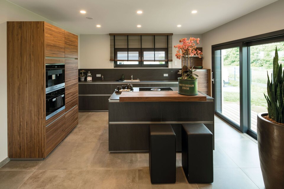Moderne Küche mit Holzfronten, Kücheninsel mit zwei schwarzen Hockern, großen Fenstern und Pflanzen im Raum des FingerHaus Fertighauses
