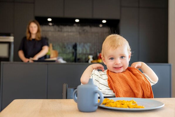 Ein Kleinkind sitzt am Esstisch eines modernen FingerHaus-Hauses mit Teller und Becher vor sich, während im Hintergrund die Mutter in der offenen Küche steht.