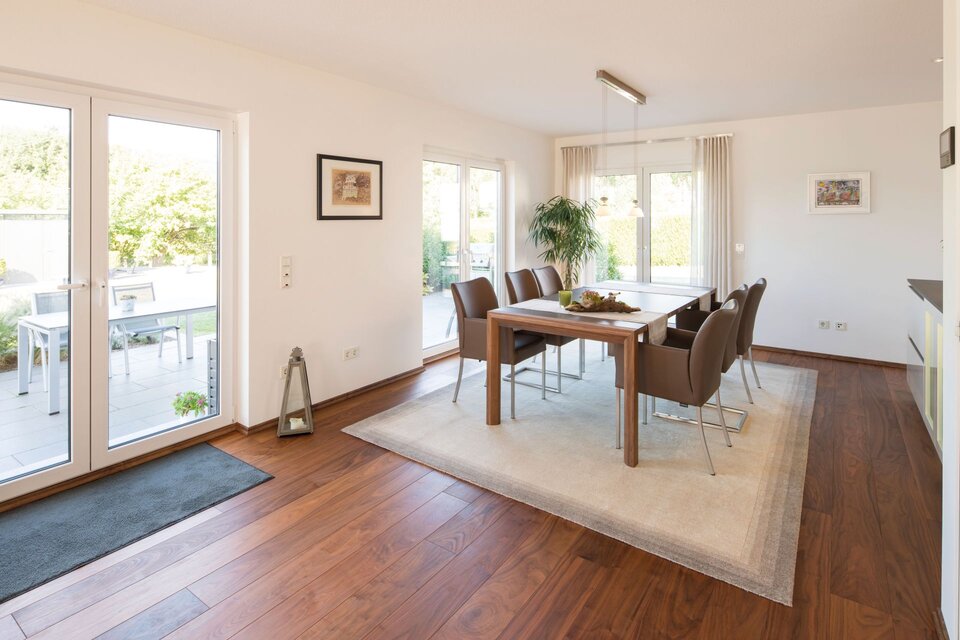 Helles Esszimmer in einem frei geplanten FingerHaus mit großem Holztisch, braunen Stühlen, hellem Teppich, bodentiefen Fenstern und Zugang zur Terrasse.