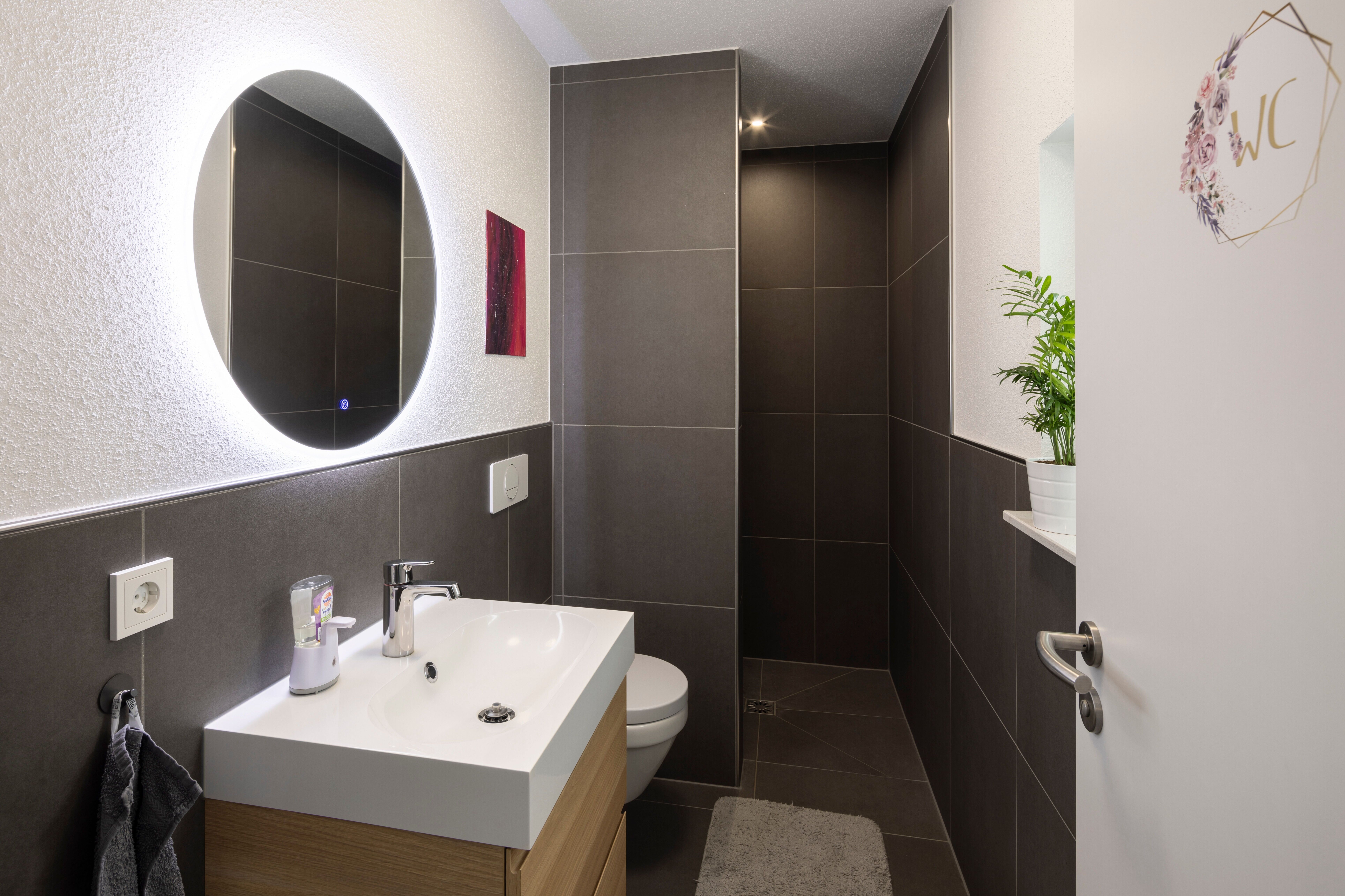 Schmaler moderner Badezimmerflur mit weißem Waschbecken auf Holzunterschrank, rundem Spiegel, Toilette und grauem Boden- und Wandfliesenbelag