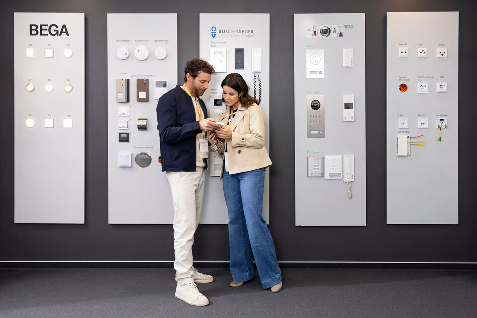 Ein Paar steht in der Bemusterung von FingerHaus vor einer Wand mit Schaltern, Steckdosen und Haustechnik-Elementen verschiedener Marken. Beide betrachten gemeinsam ein Musterstück und wirken interessiert.
