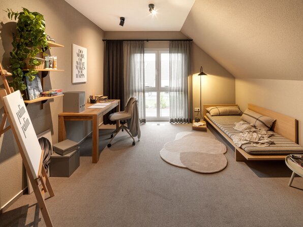 Helles Kinder- oder Jugendzimmer im Musterhaus JUNO 300 in Mülheim-Kärlich mit Schreibtisch, gemütlichem Tagesbett und dekorativen Wandregalen.