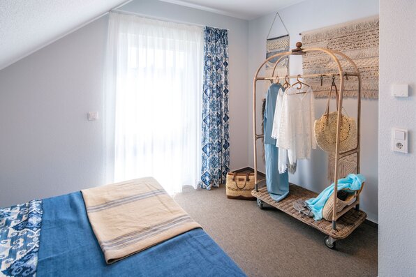Schlafzimmer im FingerHaus-Musterhaus JUNO in Frankenberg mit Doppelbett unter Dachschräge, blau-beiger Bettwäsche, bodentiefem Fenster mit transparentem Vorhang und blau gemustertem Seitenschal sowie offenem Kleiderständer aus Holz mit Kleidung und Taschen.