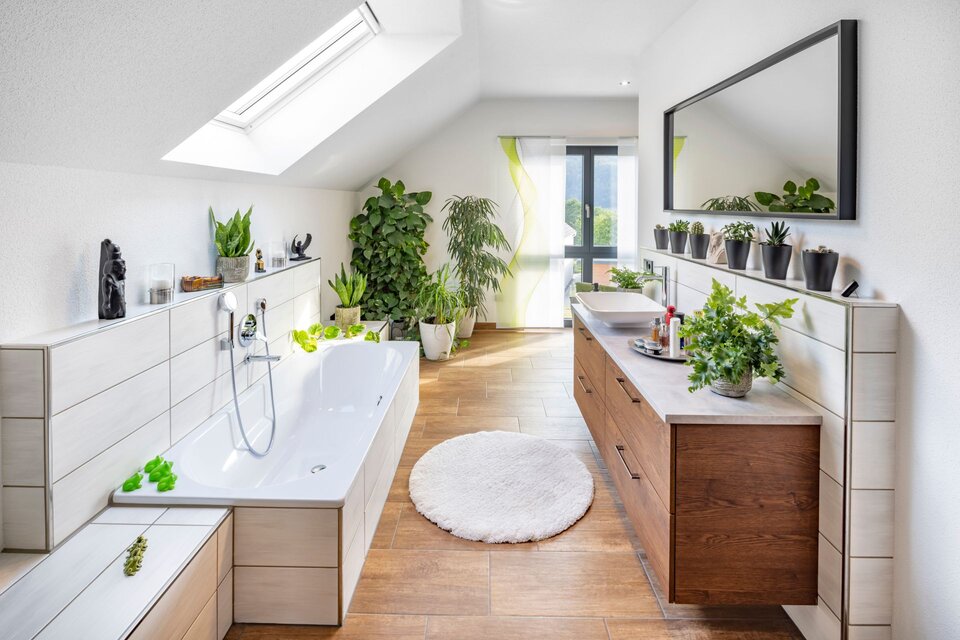 Großes Badezimmer mit Badewanne unter der Dachschräge, Holzfronten und vielen Pflanzen. Helles Tageslicht, runder Teppich und großer Spiegel schaffen eine frische, natürliche Atmosphäre.