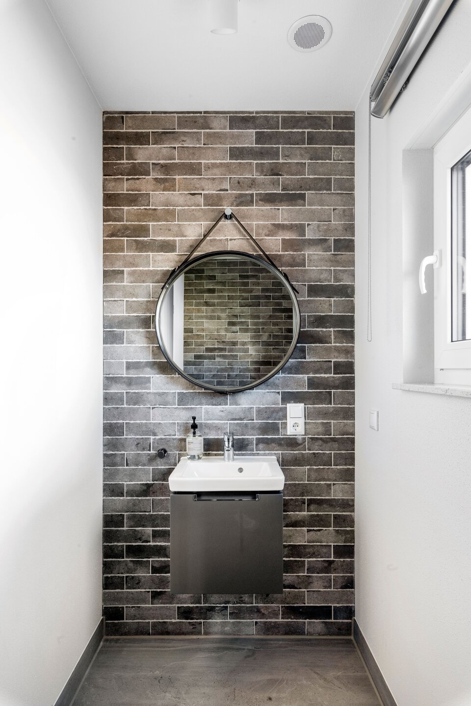Modernes Gäste-WC im FingerHaus Musterhaus TALO in Babenhausen mit grauen Backsteinfliesen, kleinem Waschbecken und rundem Spiegel mit Lederriemen. Minimalistisches Design mit klaren Linien und hellen Akzenten.