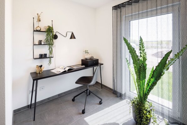 Modernes Homeoffice im Musterhaus TALO Fulda mit schmalem schwarzen Schreibtisch, Designstuhl und großem Fenster mit leichten Vorhängen, das viel Tageslicht hereinlässt.