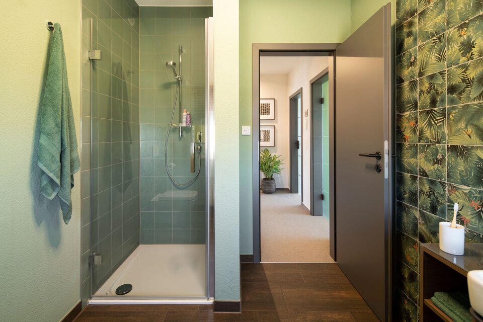 Kleines Badezimmer mit grünen Wandfliesen in der Dusche und dunklem Holzboden. Eine Wand mit tropischen Fliesen, Handtuchhalter und offener Tür mit Blick in den Flur.