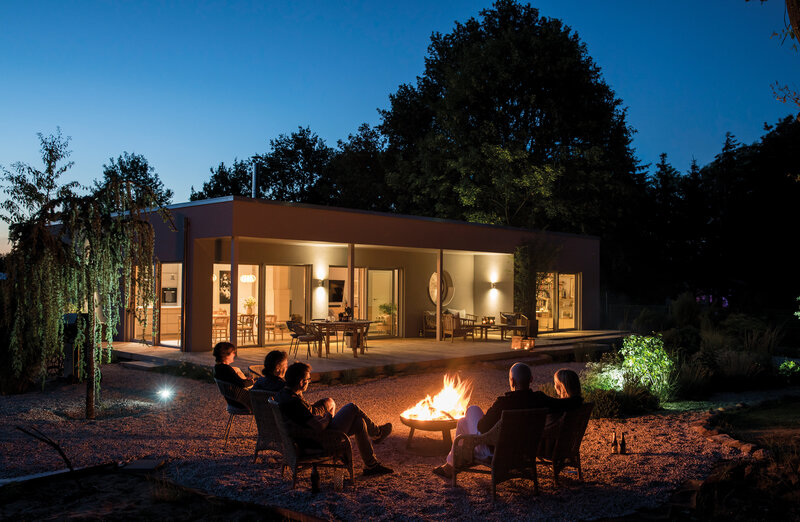 Moderner Flachdach-Bungalow NIVO bei Nacht mit beleuchteter Terrasse und Gruppe von Personen am Feuer im Garten