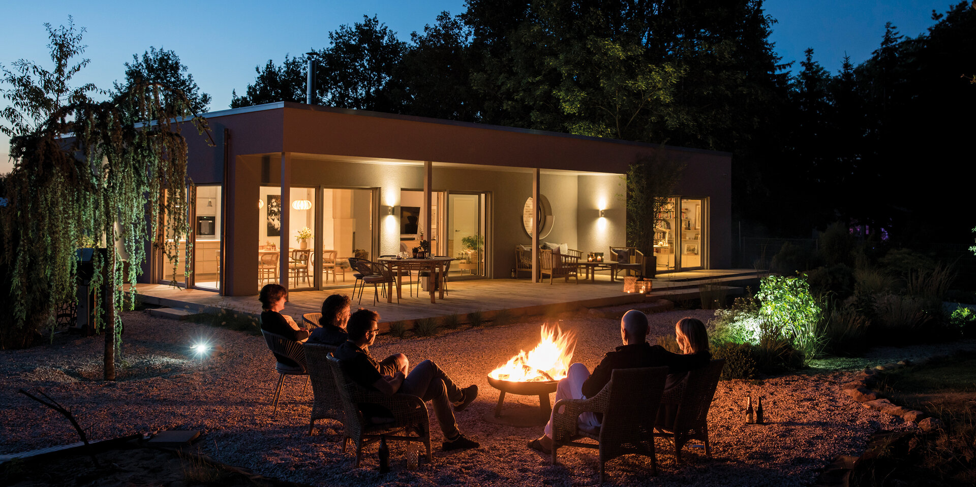 Moderner Flachdach-Bungalow NIVO bei Nacht mit beleuchteter Terrasse und Gruppe von Personen am Feuer im Garten