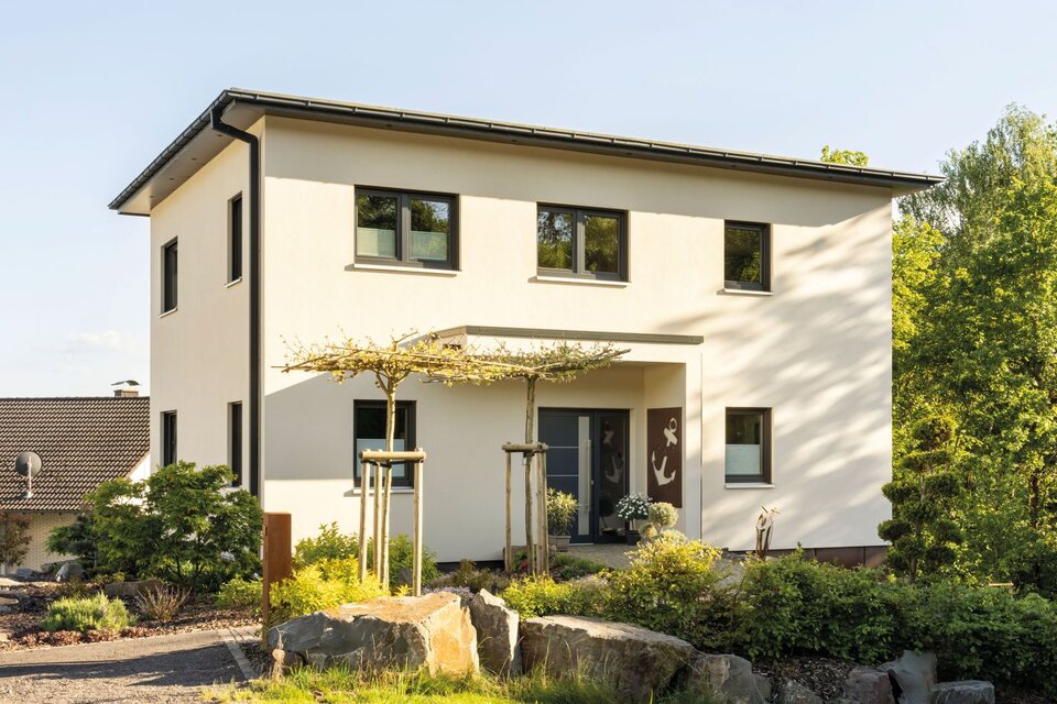 Modernes zweigeschossiges FingerHaus mit heller Fassade, Satteldach, überdachtem Eingangsbereich und gepflegtem Vorgarten mit Natursteinen und Bäumen.