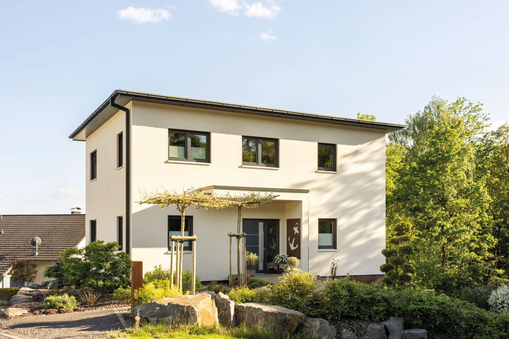 Modernes zweigeschossiges FingerHaus mit heller Fassade, Satteldach, überdachtem Eingangsbereich und gepflegtem Vorgarten mit Natursteinen und Bäumen.