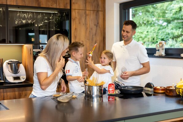 Eine Familie kocht gemeinsam in der modernen Küche eines FingerHaus-Hauses. Die Eltern und zwei Kinder lachen und rühren fröhlich in einem Topf mit Spaghetti.
