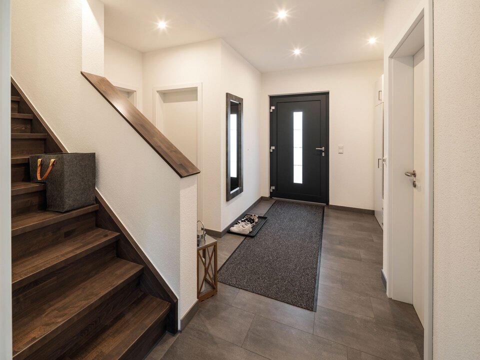 Eingangsbereich mit dunkler Holztreppe, grauem Fliesenboden und moderner Haustür mit Glaseinsätzen. Dezente Beleuchtung und helle Wände schaffen einen freundlichen Empfangsbereich.