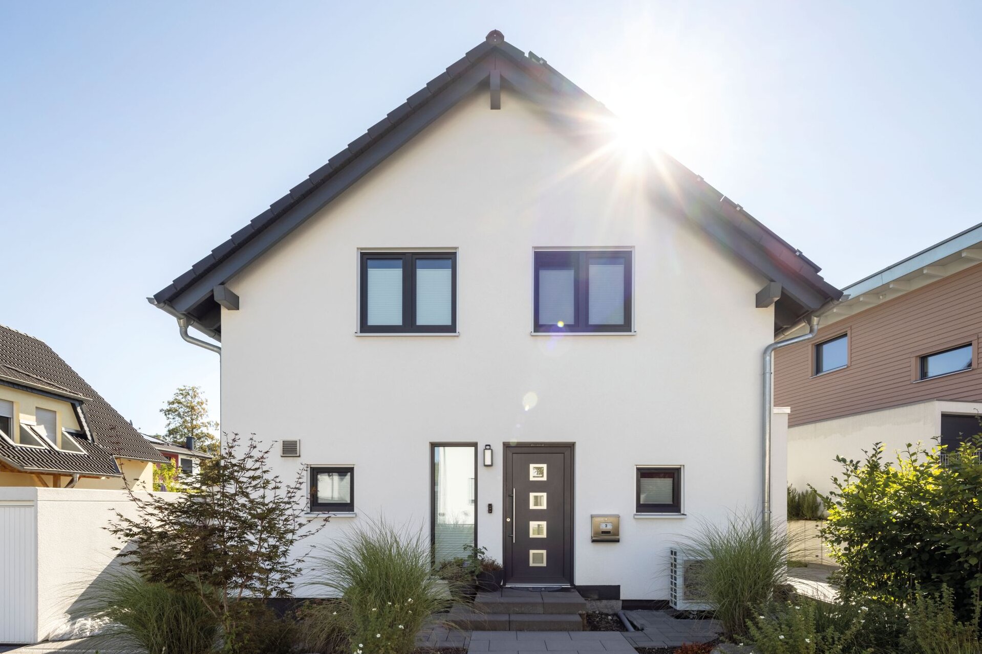 Frontansicht eines modernen Einfamilienhauses mit weißer Putzfassade, dunklem Satteldach und anthrazitfarbener Haustür. Ein gepflasterter Weg führt durch den Vorgarten mit Ziergräsern und Steinen zum Eingang.