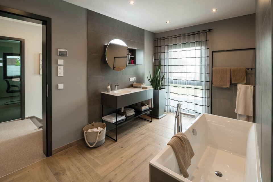 Modernes Badezimmer im Musterhaus ALTERO in Gießen mit rechteckiger Badewanne, Waschtisch und großer Fensterfront mit Vorhängen.
