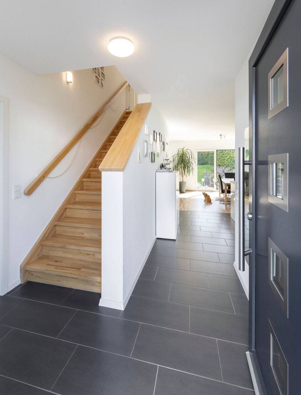Flur mit geschlossener Holztreppe, weißen Wänden und dunklem Boden. Im Hintergrund Wohnbereich mit hellen Möbeln, Tageslicht durch Fenster.