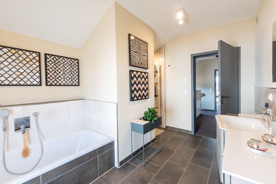 Modernes Badezimmer im FingerHaus Musterhaus TALO in Babenhausen mit Badewanne, Doppelwaschbecken und dekorativen Wandbildern. Helle Wandgestaltung kombiniert mit dunklem Boden und stilvollen Einrichtungselementen.